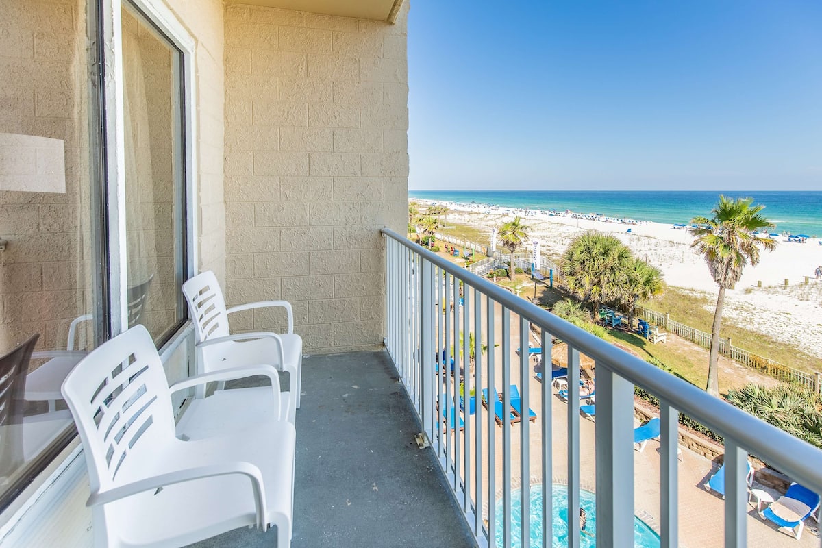A balcony with white plastic chairs, a glass table, and a view of a beach with palm trees and lounge chairs.