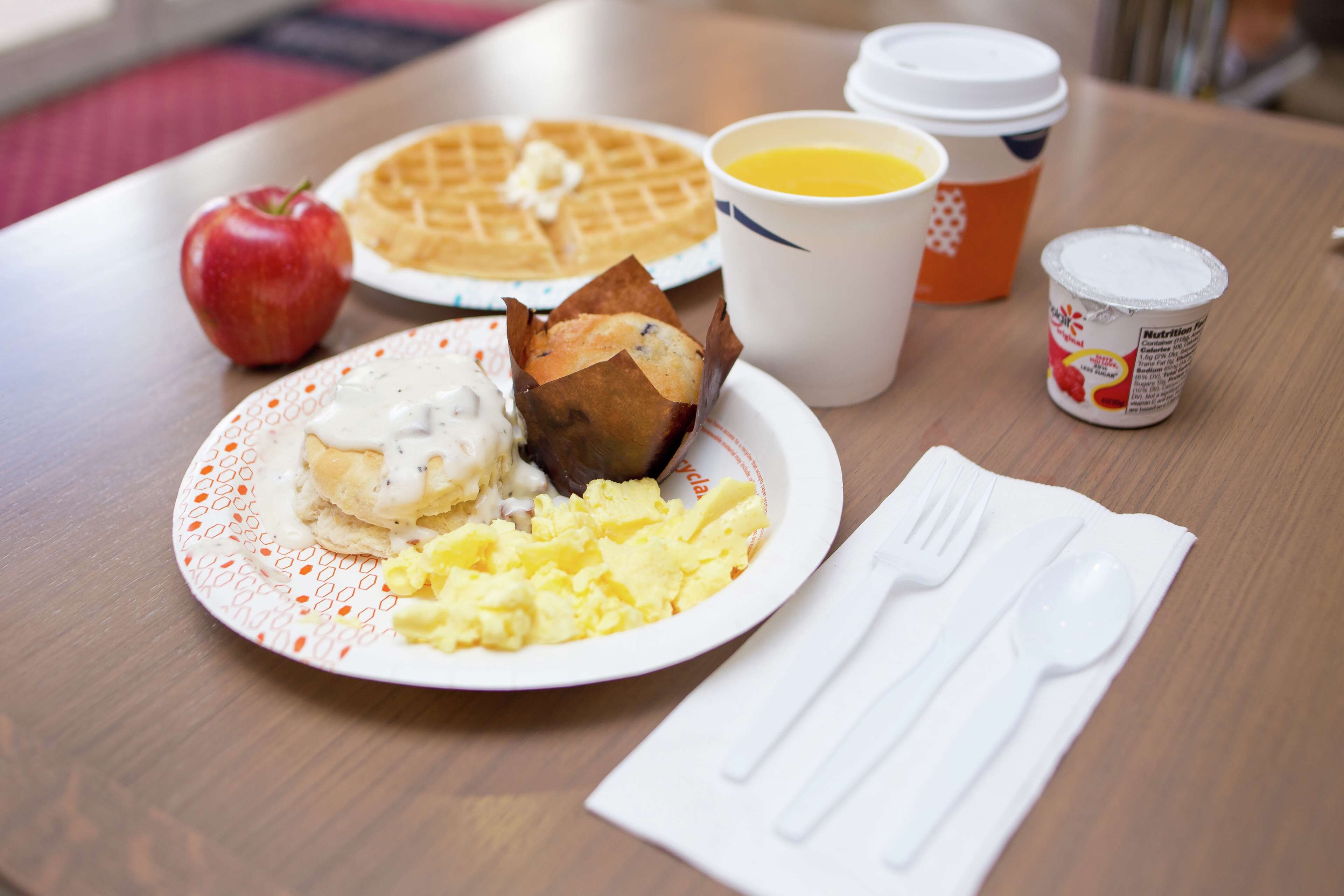 Une assiette de petit-déjeuner comprenant des œufs brouillés, un muffin et une gaufre, accompagnée d’un verre de jus d’orange et d’un pot de yaourt.