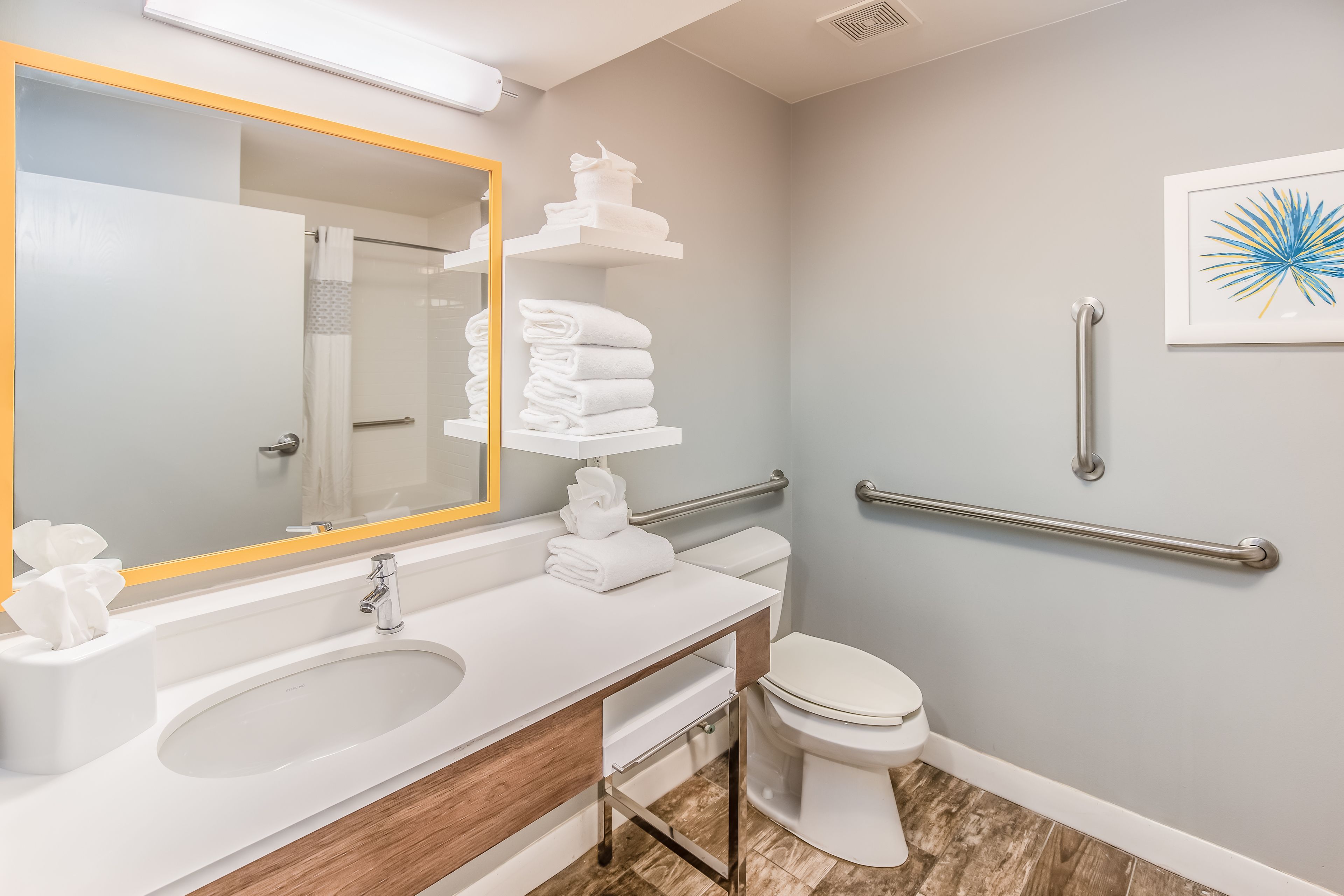 A modern bathroom with a white sink, mirror, and toilet. There are white towels stacked on a shelf, and a handrail is installed on the wall.