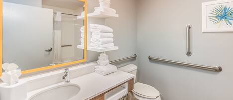 A modern bathroom with a white sink, mirror, and toilet. There are white towels stacked on a shelf, and a handrail is installed on the wall.