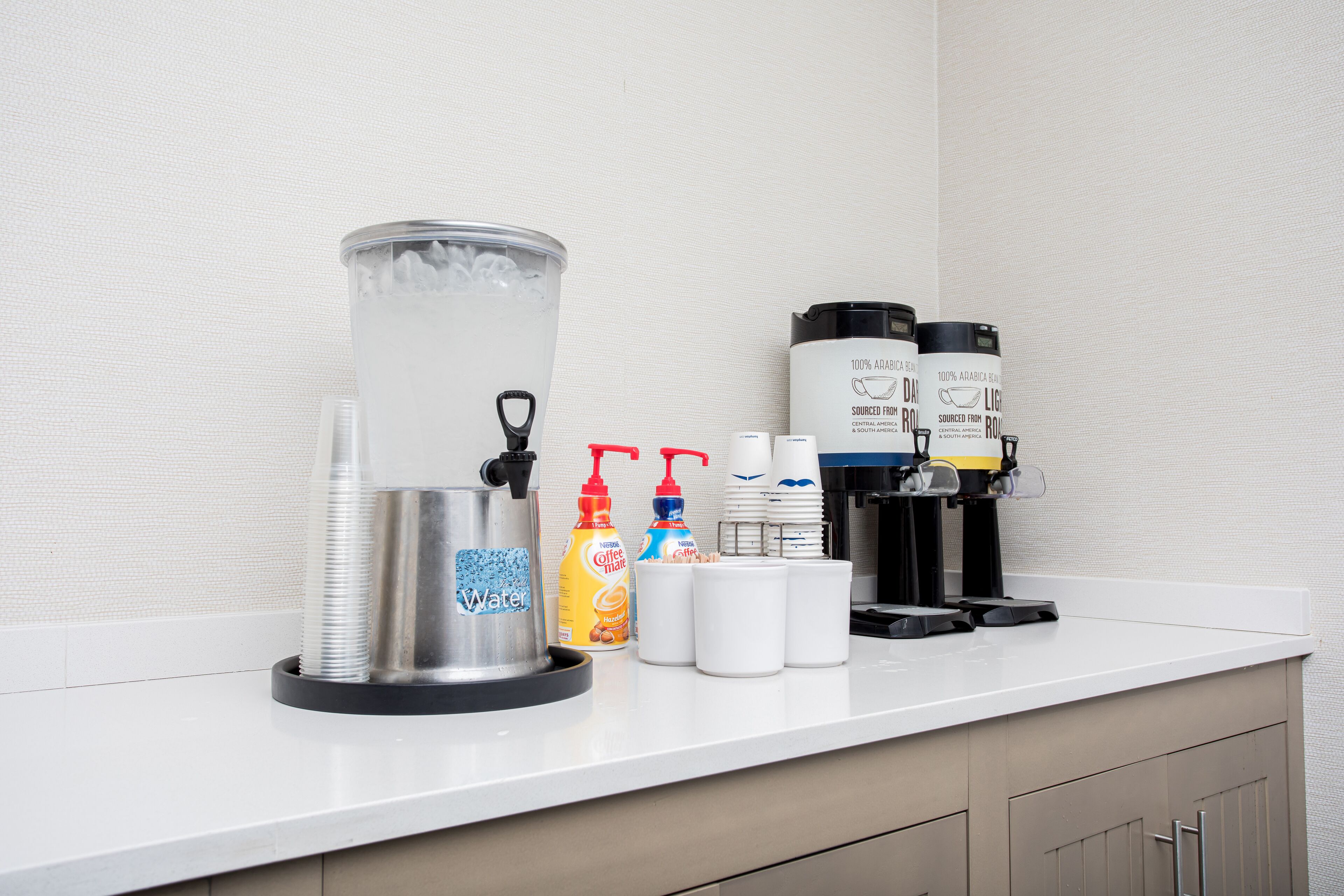 A coffee station with a water dispenser, coffee machines, and various coffee pods on a white countertop.