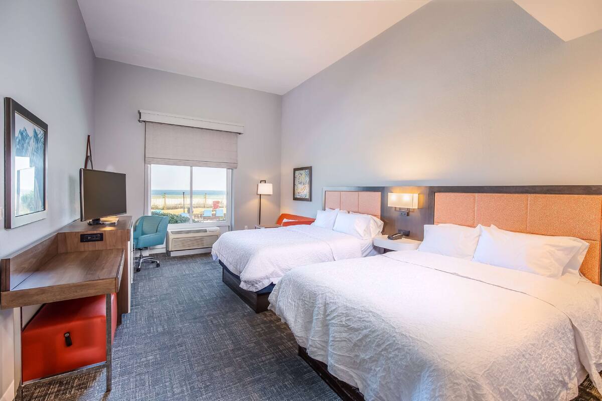 A hotel room with two beds, a desk, a chair, a TV, and a window with a view.