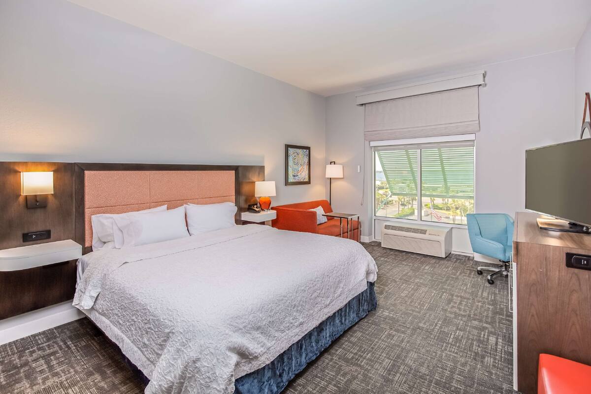 A hotel room with a large bed, a desk, a chair, a TV, and a window with blinds.