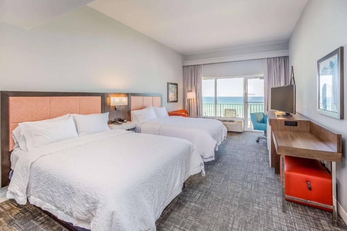 A hotel room with two beds, a desk, a TV, and a balcony with a view.