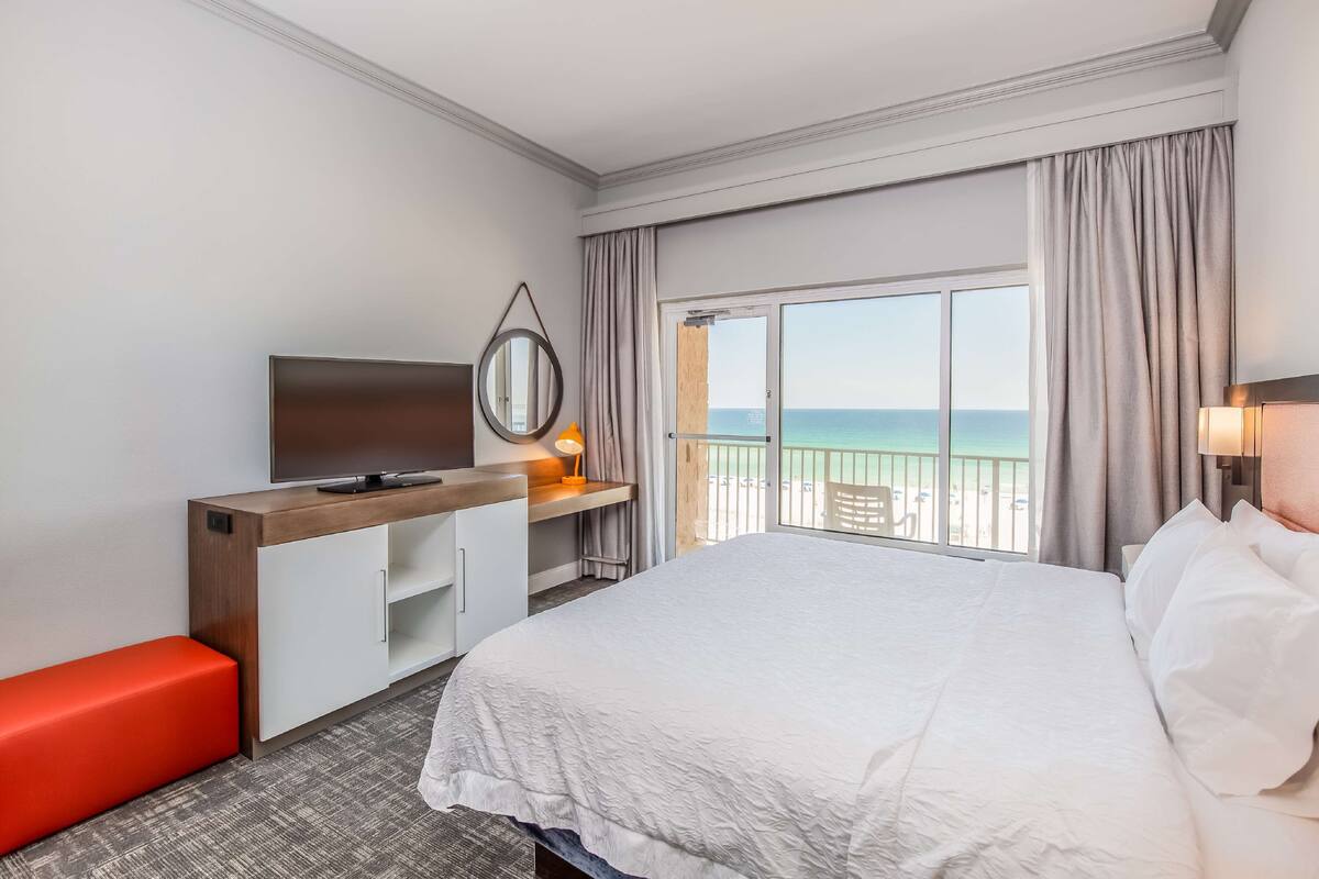 A hotel room with a large bed, a TV, a desk with a computer, a red bench, and a balcony with a view of the sea.