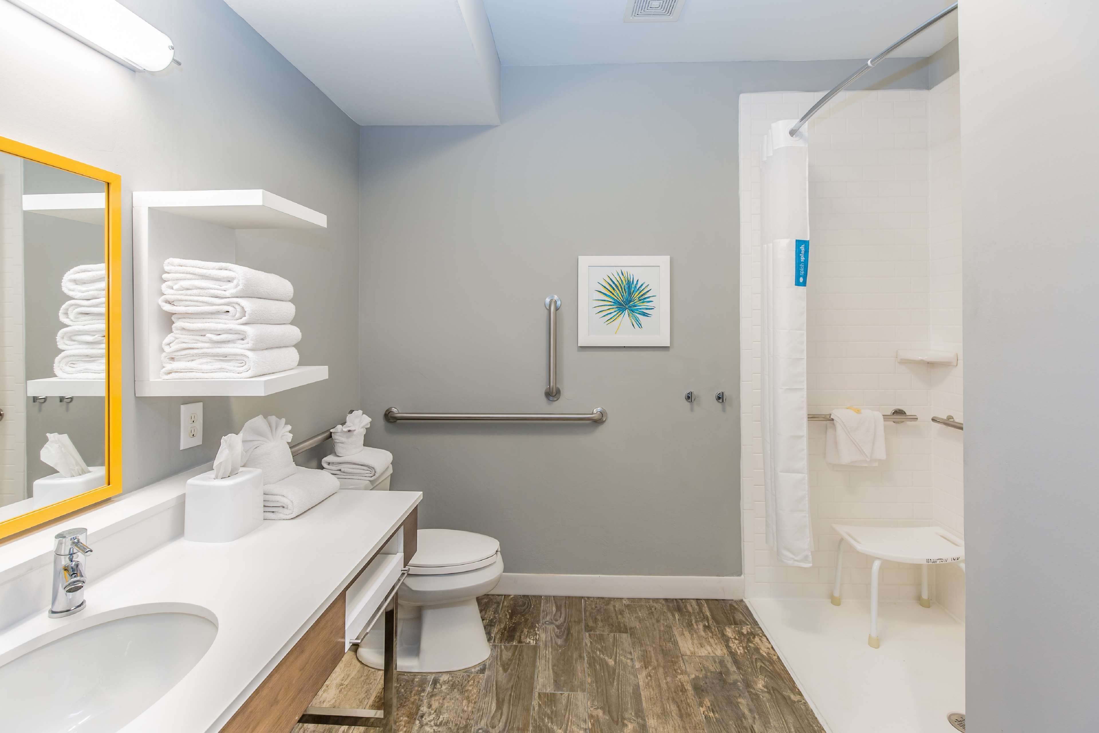 Un baño moderno con lavamanos, inodoro y zona de ducha blancos. Cuenta con un pasamanos para accesibilidad, un perchero y una obra de arte enmarcada en la pared.