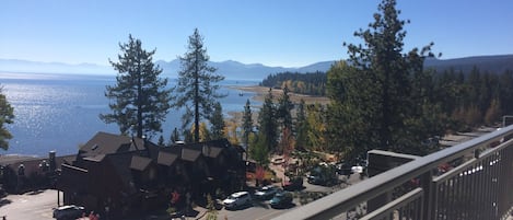 A view from a balcony overlooking a parking area with cars, a building with a sloped roof, and a body of water with mountains in the distance.