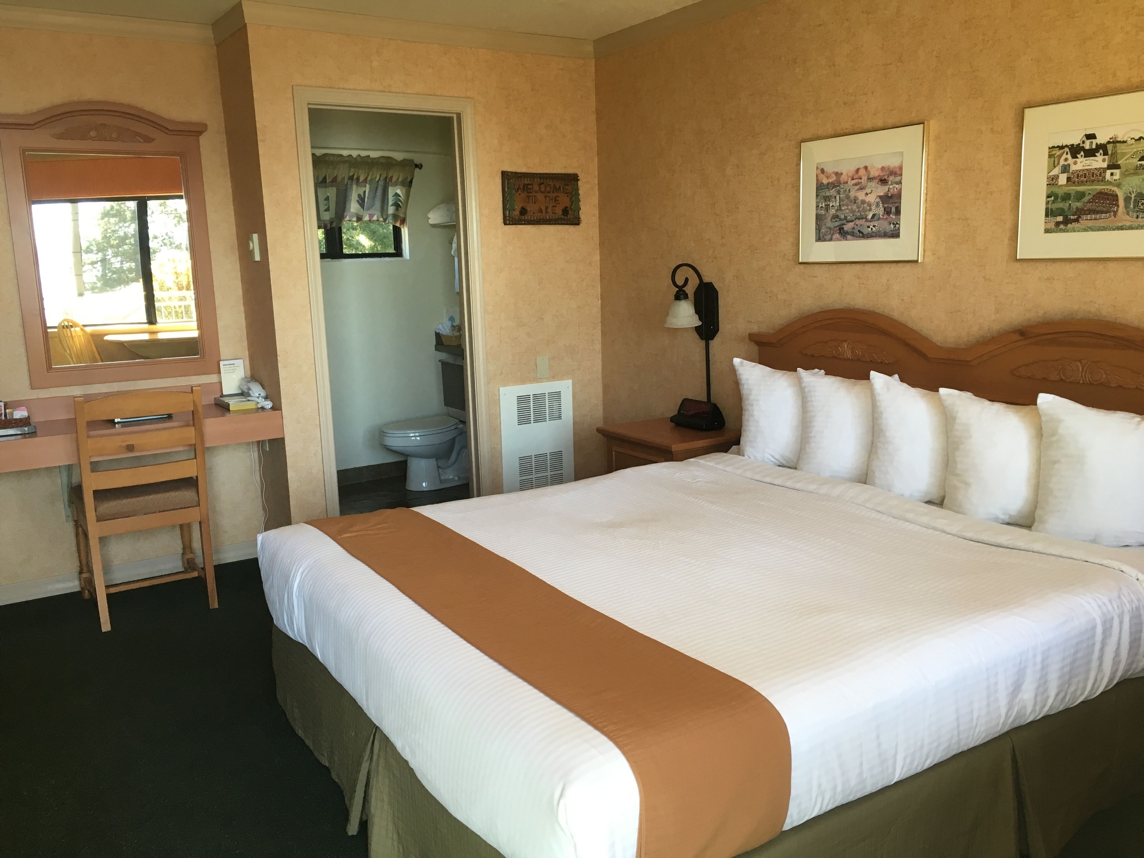 A hotel room with a bed, a desk, a chair, a toilet, and a nightstand with a lamp.