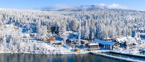 A winter scene with a lake, snow-covered buildings, and a forest in the background.