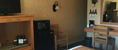 A hotel room with a bed, a desk with a chair, a microwave, a small refrigerator, a television, and a wall-mounted clock.