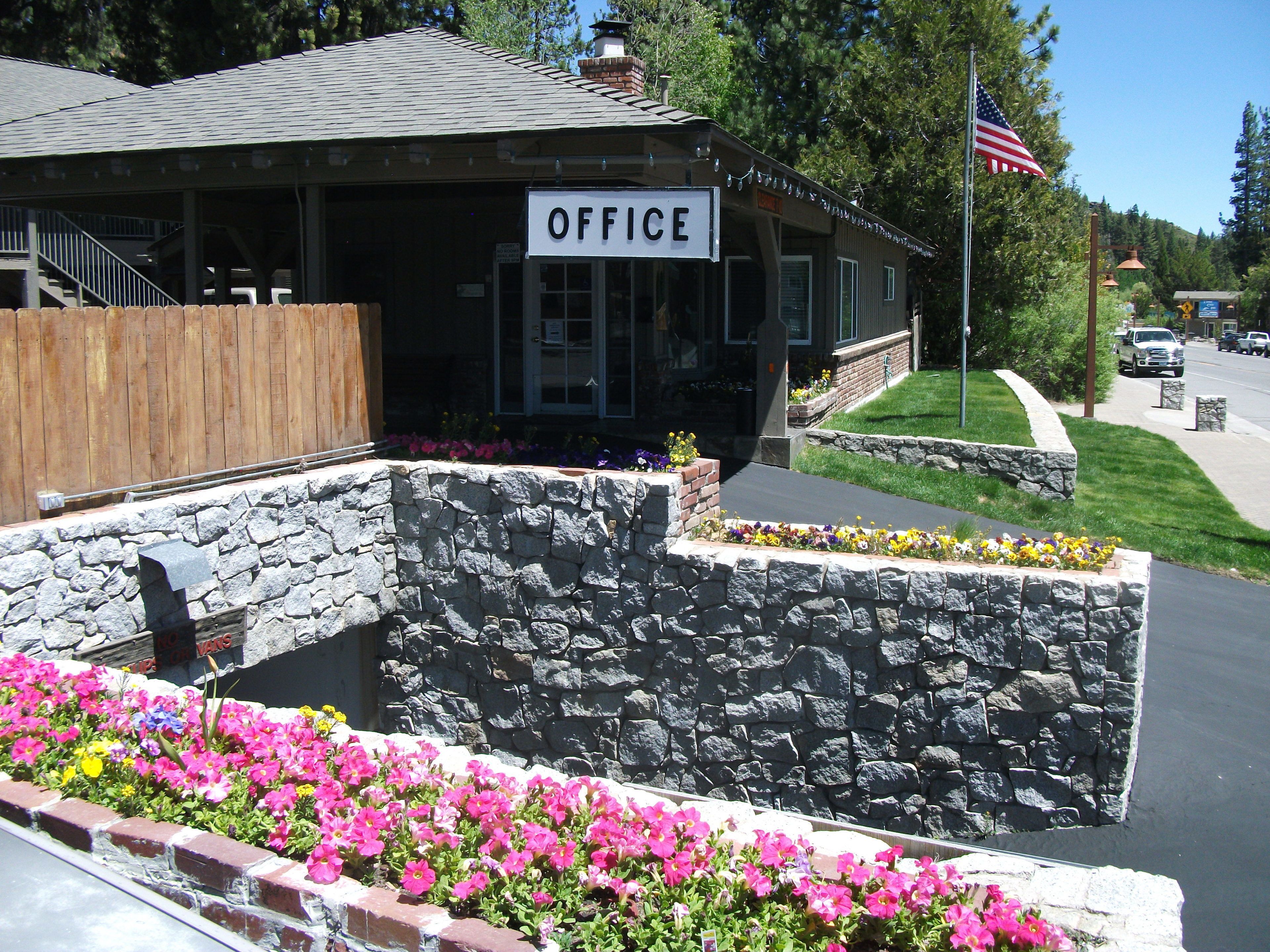A building with a sign that reads "OFFICE" and a stone wall with flowers in front.