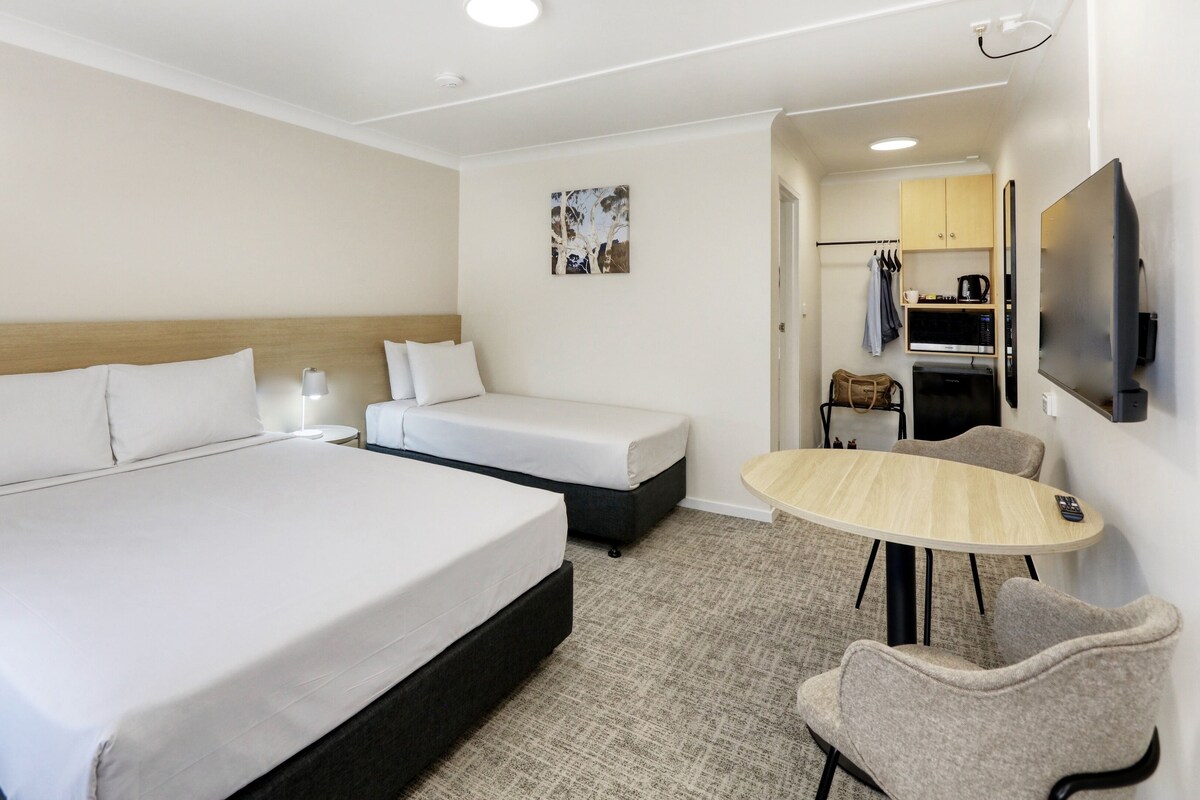 Habitación de hotel con una cama grande, una cama más pequeña, un escritorio, una silla, un televisor y un perchero con ropa.