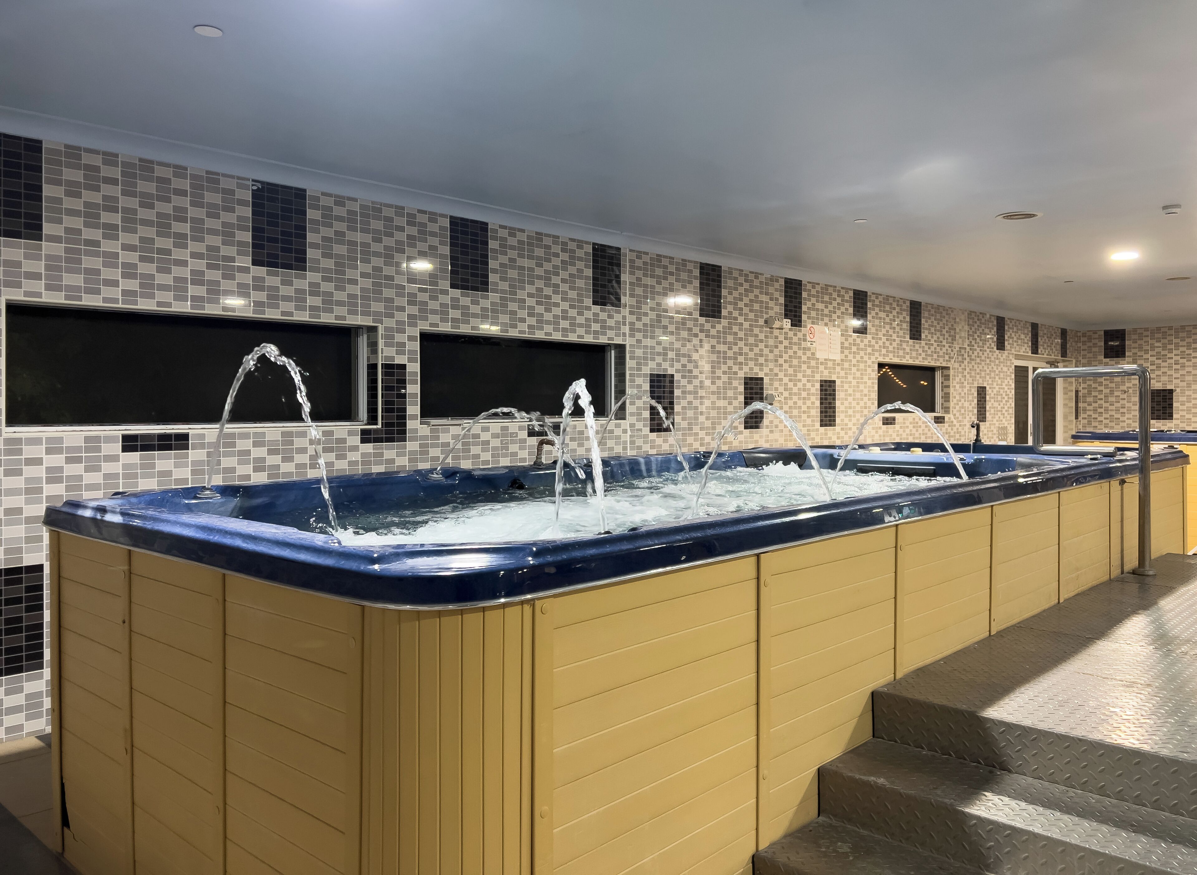 Ein großes Innenwhirlpool mit zahlreichen Wasserstrahldüsen, vor einer gefliesten Wand mit Fenstern.