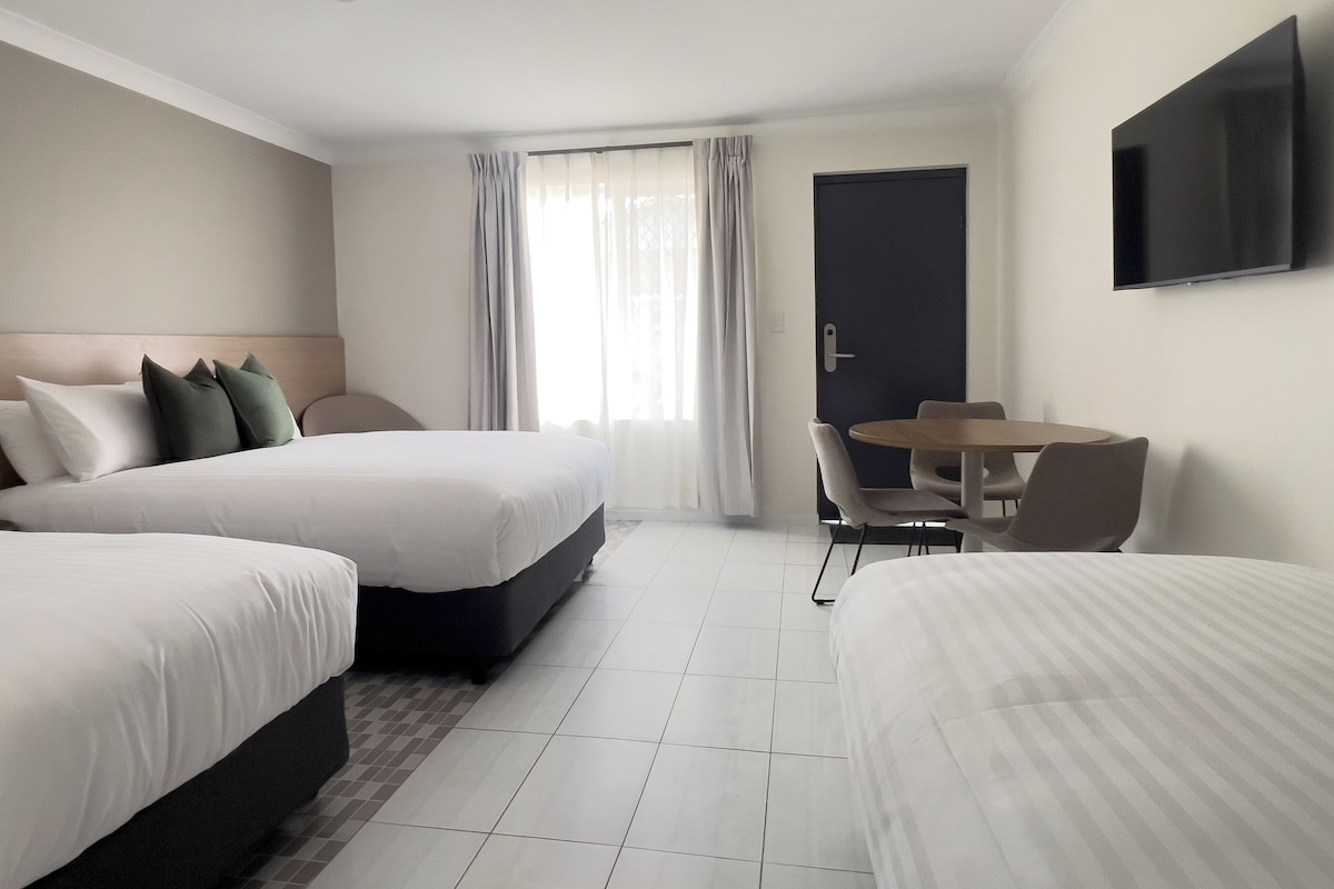 Una habitación de hotel con dos camas, una mesa de comedor con sillas, un televisor montado en la pared y una puerta con picaporte.