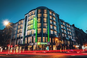 Ein mehrgeschossiges Hotel mit geschwungener Fassade, beleuchteter Beschilderung und Straßenszene mit Lichtspuren.