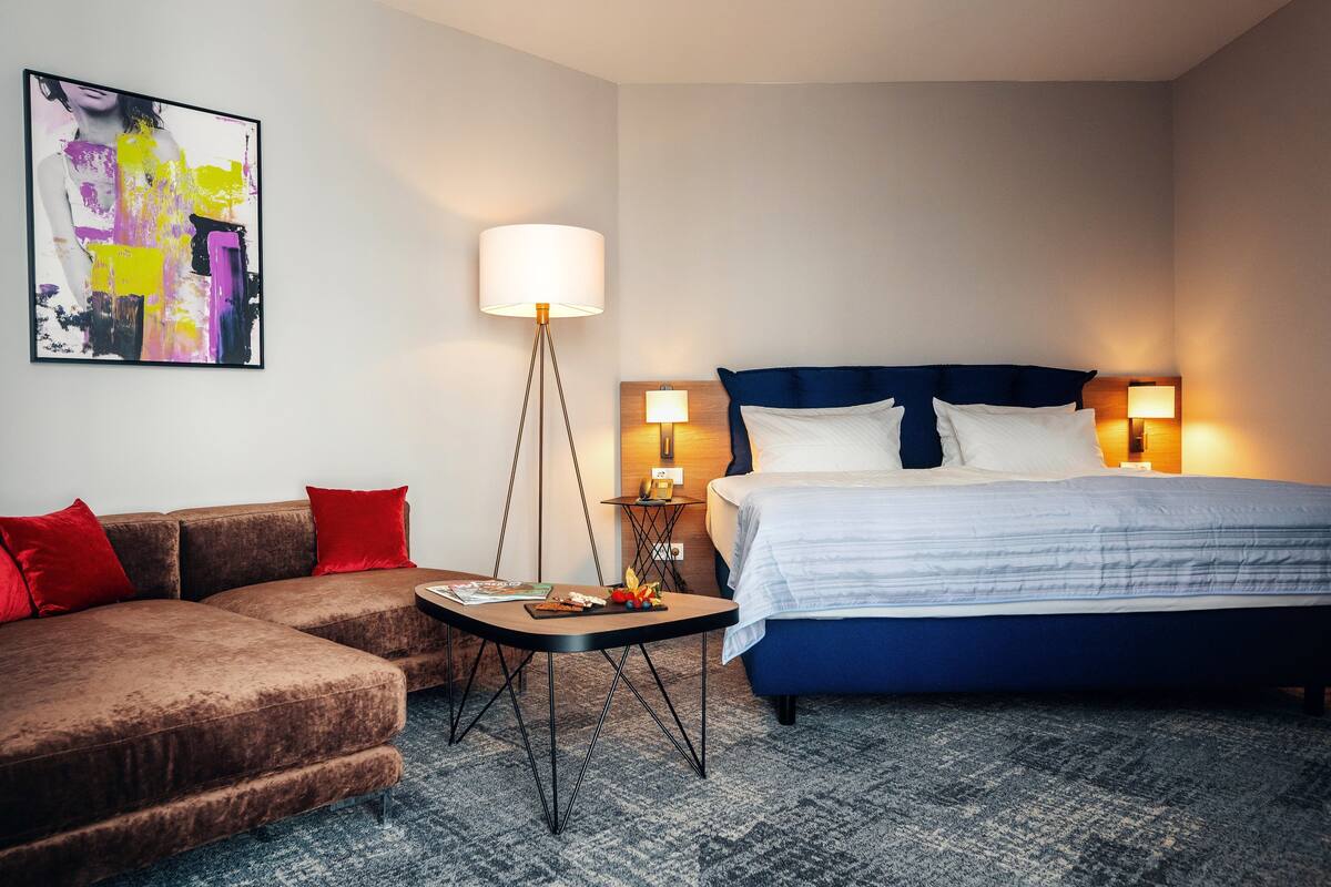 A hotel room with a bed, a sofa, a small table, a lamp, and a painting on the wall.