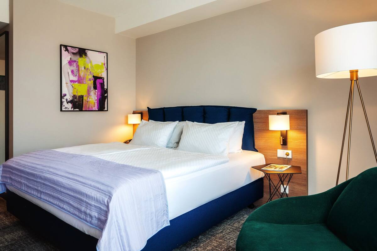 A hotel room with a large bed, two bedside lamps, a small table with a lamp, a green armchair, and a colorful abstract painting on the wall.