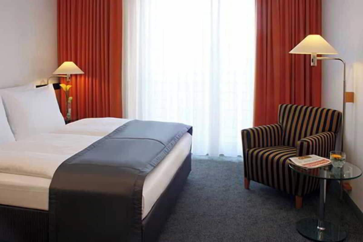 A hotel room with a bed, two lamps, a striped armchair, a small table with a magazine, and a window with curtains.