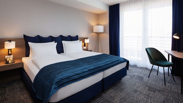 Ein Hotelzimmer mit einem großen Bett, Nachttischlampen, einem Schreibtisch mit Stuhl und einem Fenster mit Vorhängen.