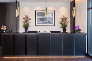 A reception desk with a framed artwork above, two potted plants, and a modern light fixture. - Holiday Inn Berlin City Center East Prenzlauer Berg by IHG (Berlin)