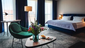 Ein Hotelzimmer mit einem Bett, einem grünen Sessel, einem kleinen Tisch mit Blumenvase und einem Esstisch mit Speisen.