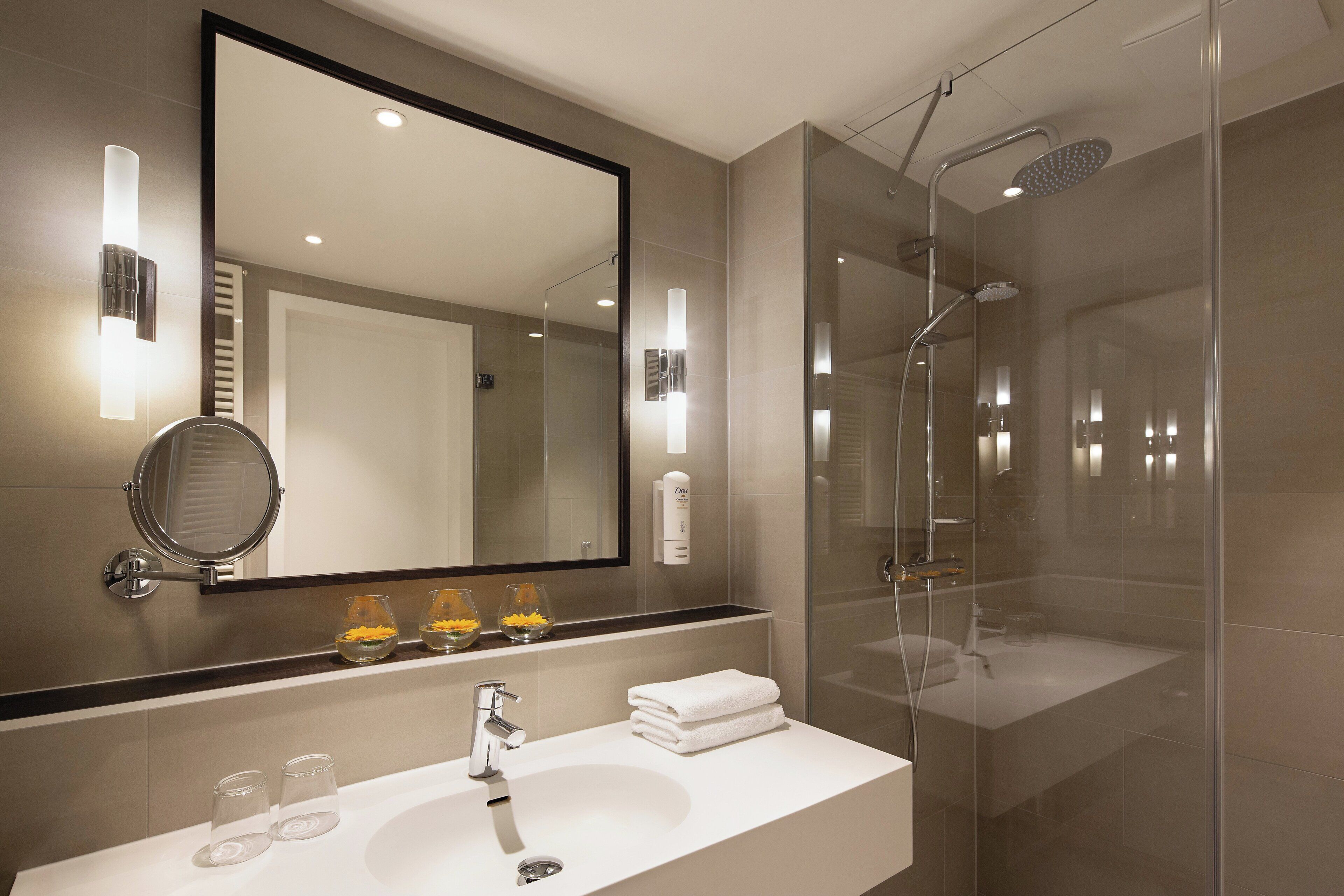 A modern bathroom with a glass-enclosed shower, a large mirror, and a vanity with a sink and two glass containers.