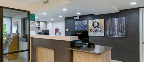 Un mostrador de recepción de hotel con un quiosco digital, un mostrador de atención al cliente y un cartel en la pared que dice 'Quality by Choice Hotels'.