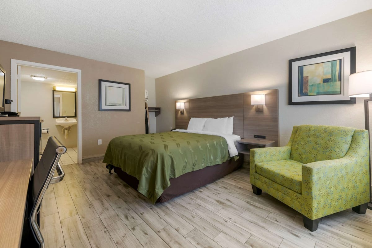 A hotel room with a bed, a green armchair, a desk, and a nightstand.