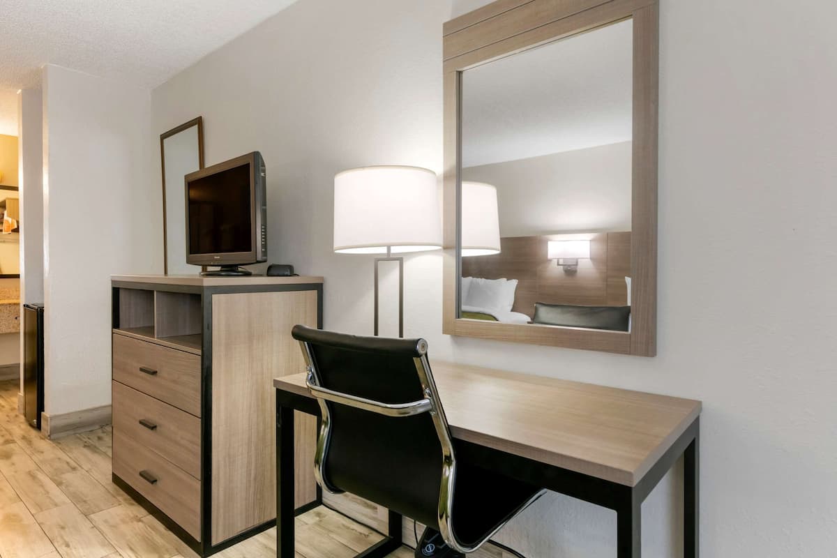A hotel room with a wooden desk, a black chair, a television, a lamp, and a mirror.