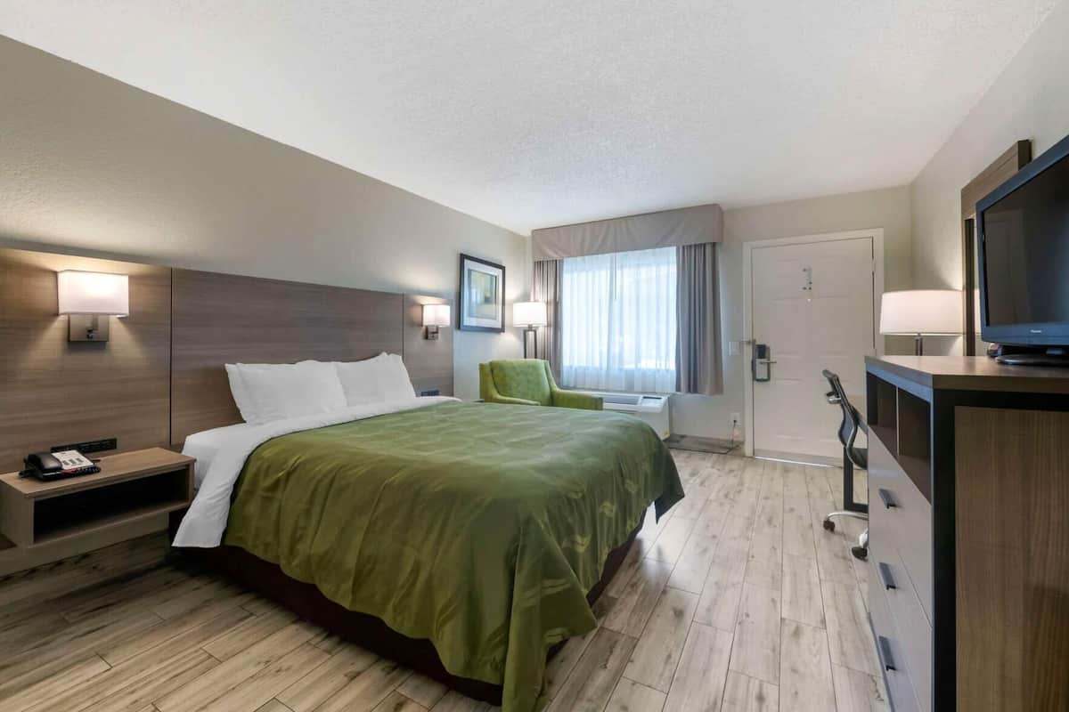 A hotel room with a bed, bedside table, lamp, chair, TV, and a desk with drawers.
