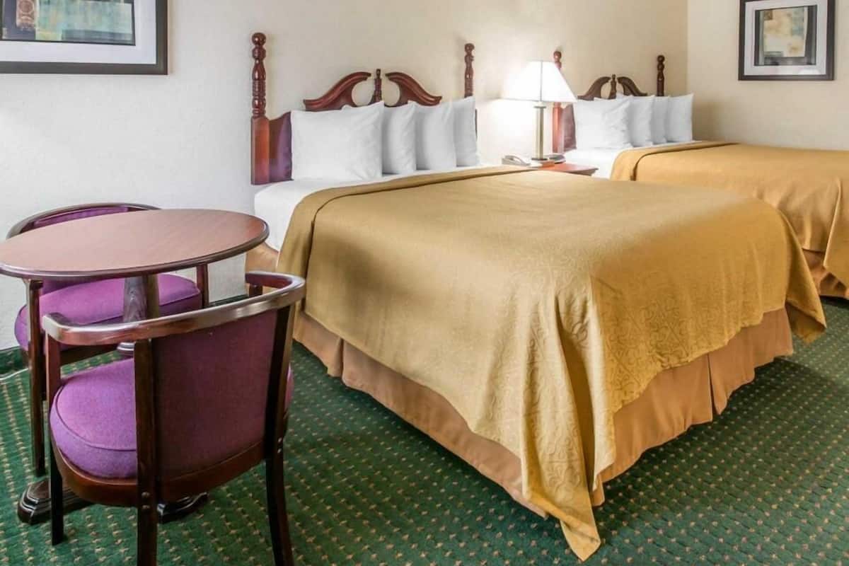 A hotel room with two beds, a small table with a chair, a lamp, and artwork on the walls.