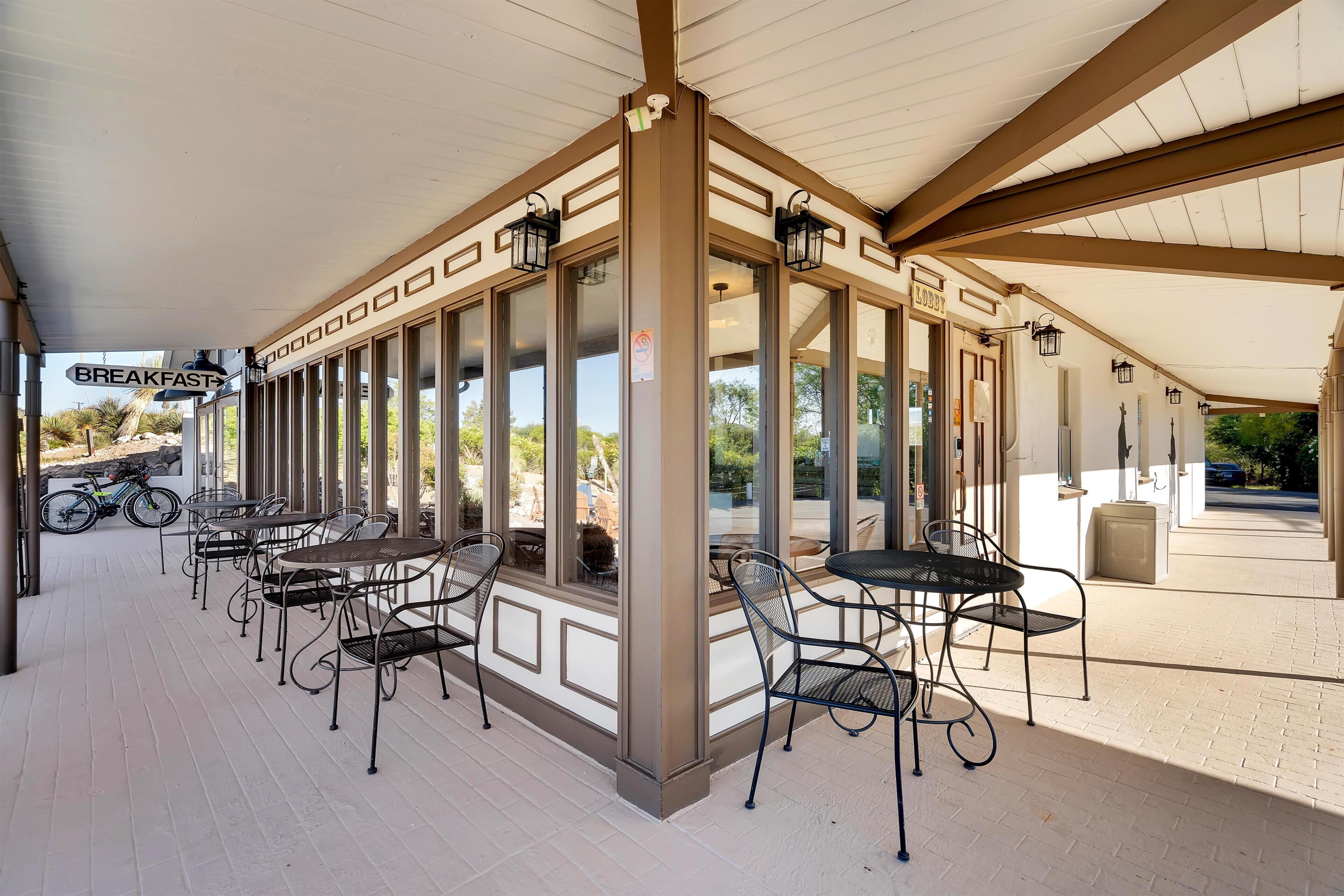 A covered outdoor seating area with tables and chairs, a sign for breakfast, and a view of the surrounding landscape.