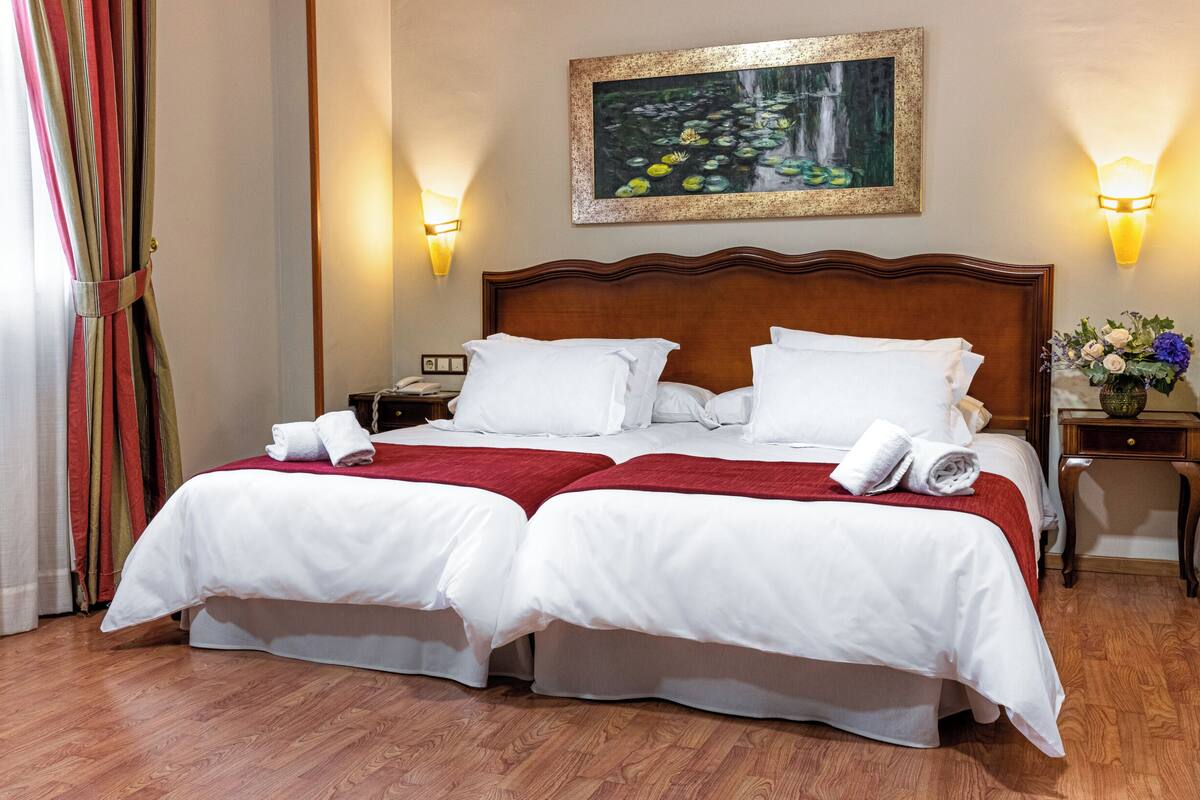 Una cama doble con sábanas blancas, una manta roja y dos toallas enrolladas. Hay una mesita de noche con una lámpara y un teléfono, y un cuadro sobre la cama.