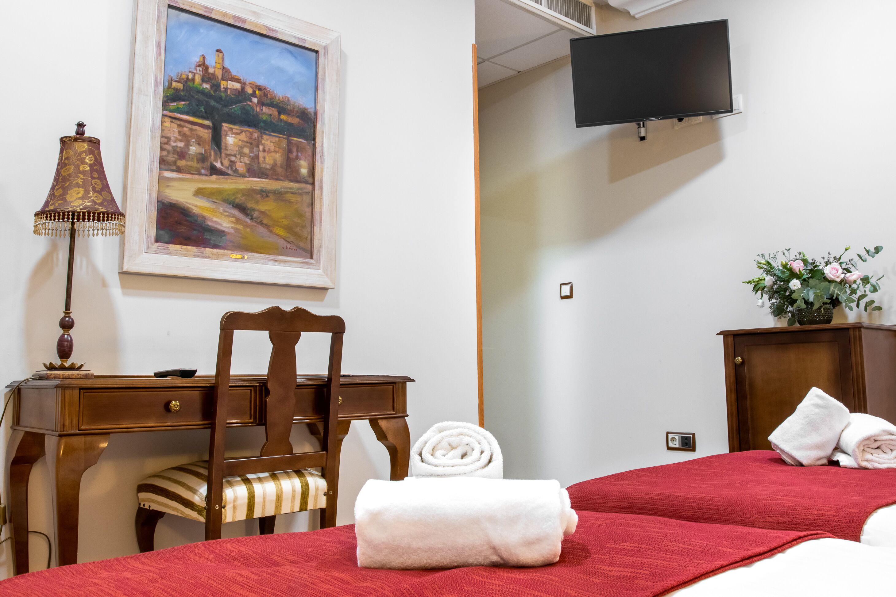 A hotel room with two beds, a desk, a chair, a painting, a TV, and a cabinet with towels.