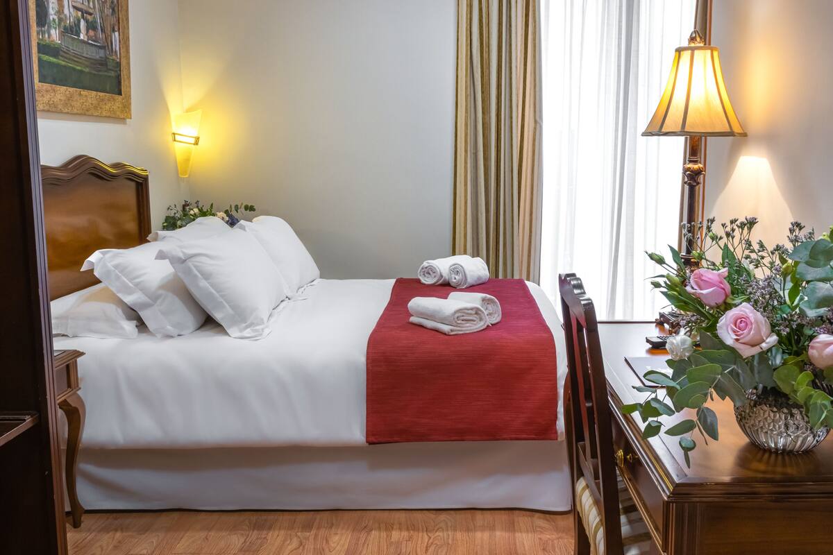 Habitación de hotel con una cama bien hecha, una manta roja y un jarrón con flores sobre una mesita de noche.