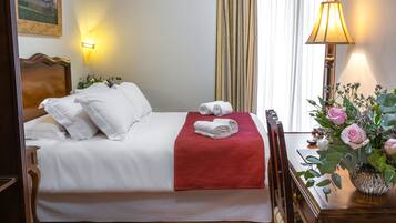 Une chambre d’hôtel avec un lit soigneusement fait, un plaid rouge et un vase de fleurs posé sur une table de chevet en bois.