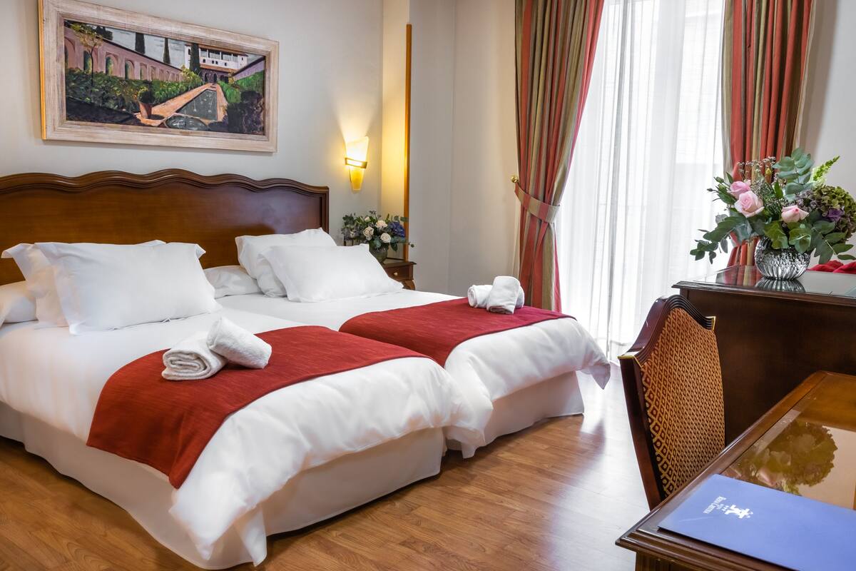 Habitación de hotel con dos camas, cabecera de madera, escritorio con silla, un cuadro en la pared y una ventana con cortinas.