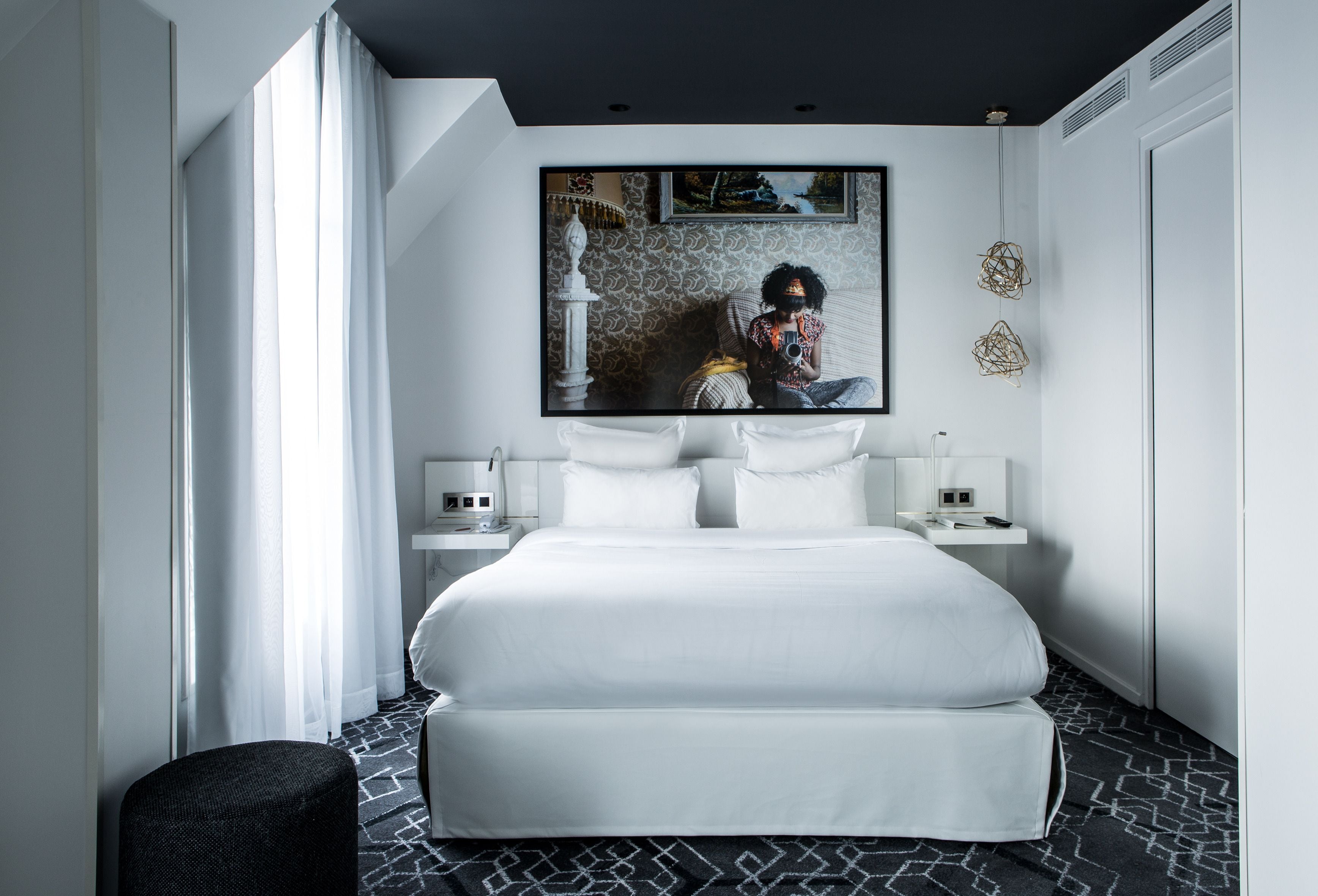 A modern hotel room with a large bed, a flat-screen TV mounted on the wall, a bedside table with a lamp, and a patterned floor.
