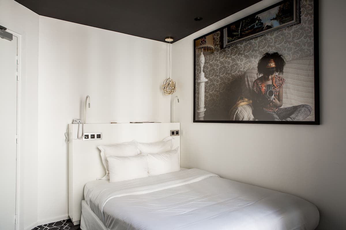 A modern bedroom with a white bed, a headboard with controls, a wall-mounted lamp, and a framed picture.