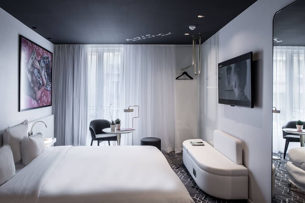 A modern hotel room with a large bed, a seating area, a TV, and a window with sheer curtains.