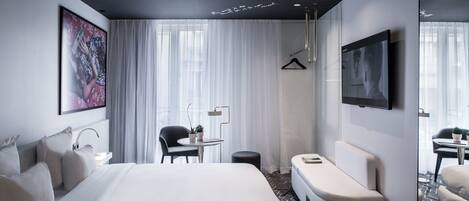 A modern hotel room with a large bed, a seating area, a TV, and a window with sheer curtains.
