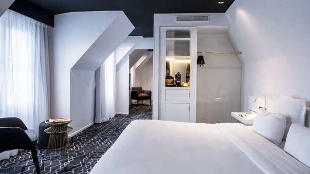 A modern hotel room with a large bed, a seating area with a chair and a small table, a walk-in closet, and a bathroom with a mirror and sink. - Le General Hotel (Paris)