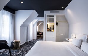 Moderni hotellihuone, jossa on suuri sänky, oleskelualue tuolilla ja pienellä pöydällä, erillinen vaatehuone sekä kylpyhuone peilillä ja pesualtaalla.