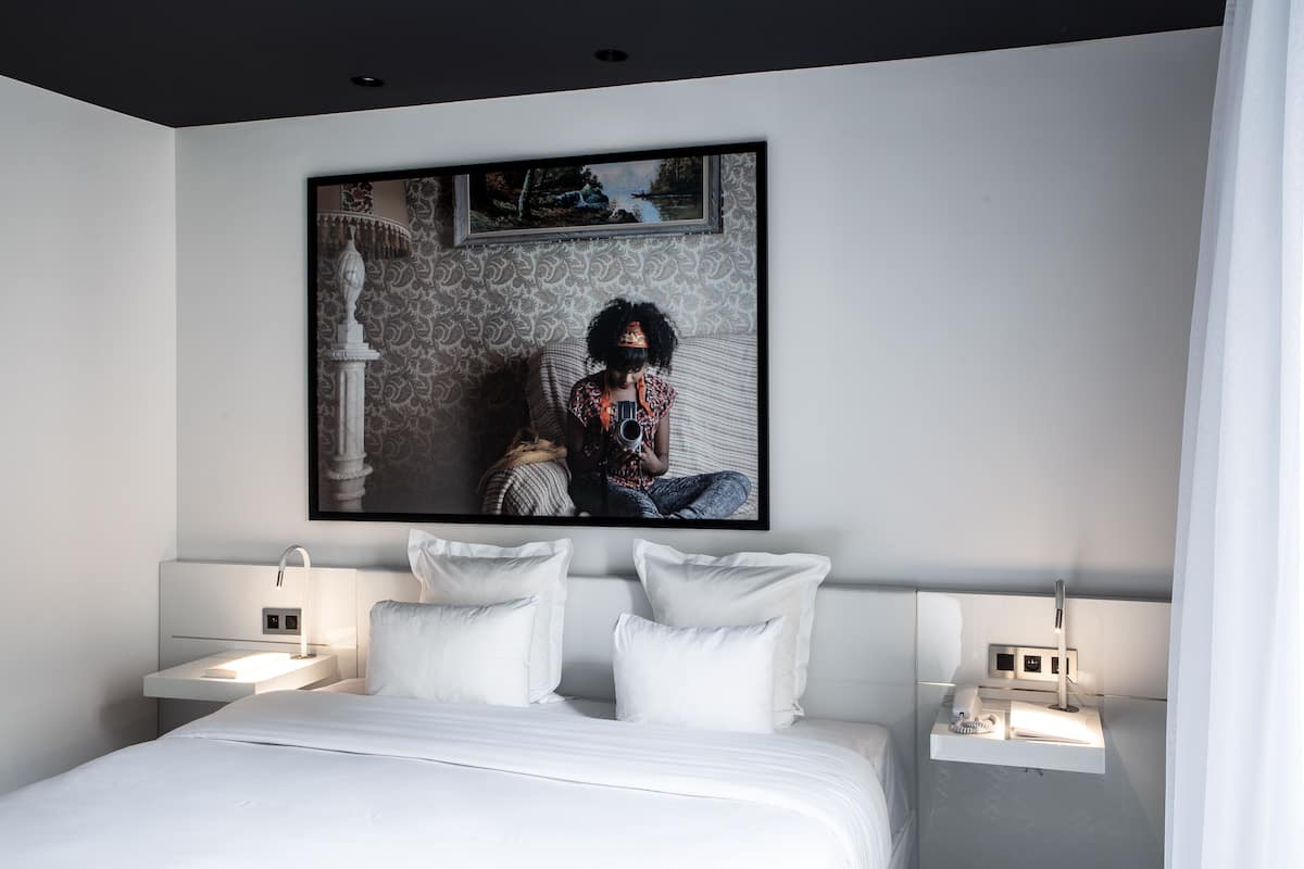 A modern bedroom with a large bed, two bedside tables, and a wall-mounted artwork depicting a person sitting by a river.