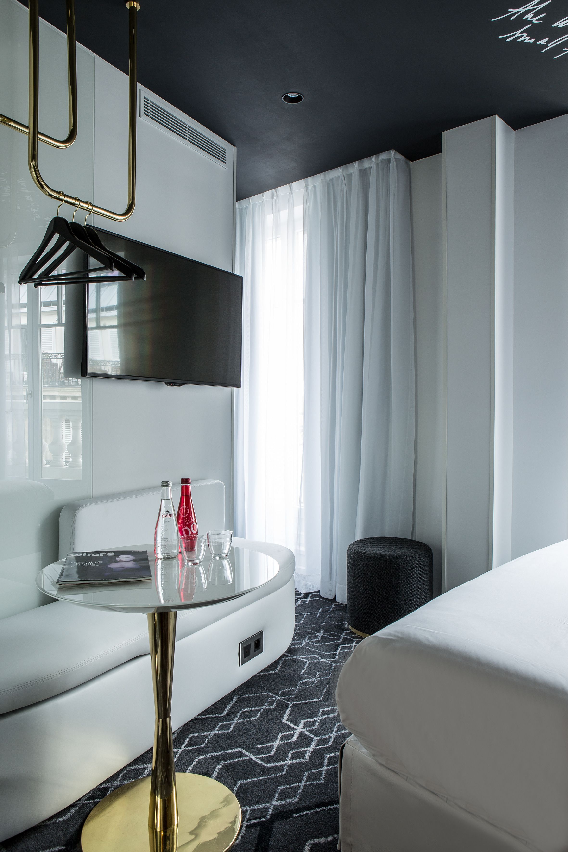 A modern hotel room with a white sofa, a glass table with a gold base, a flat-screen TV mounted on the wall, and a large window with sheer curtains.