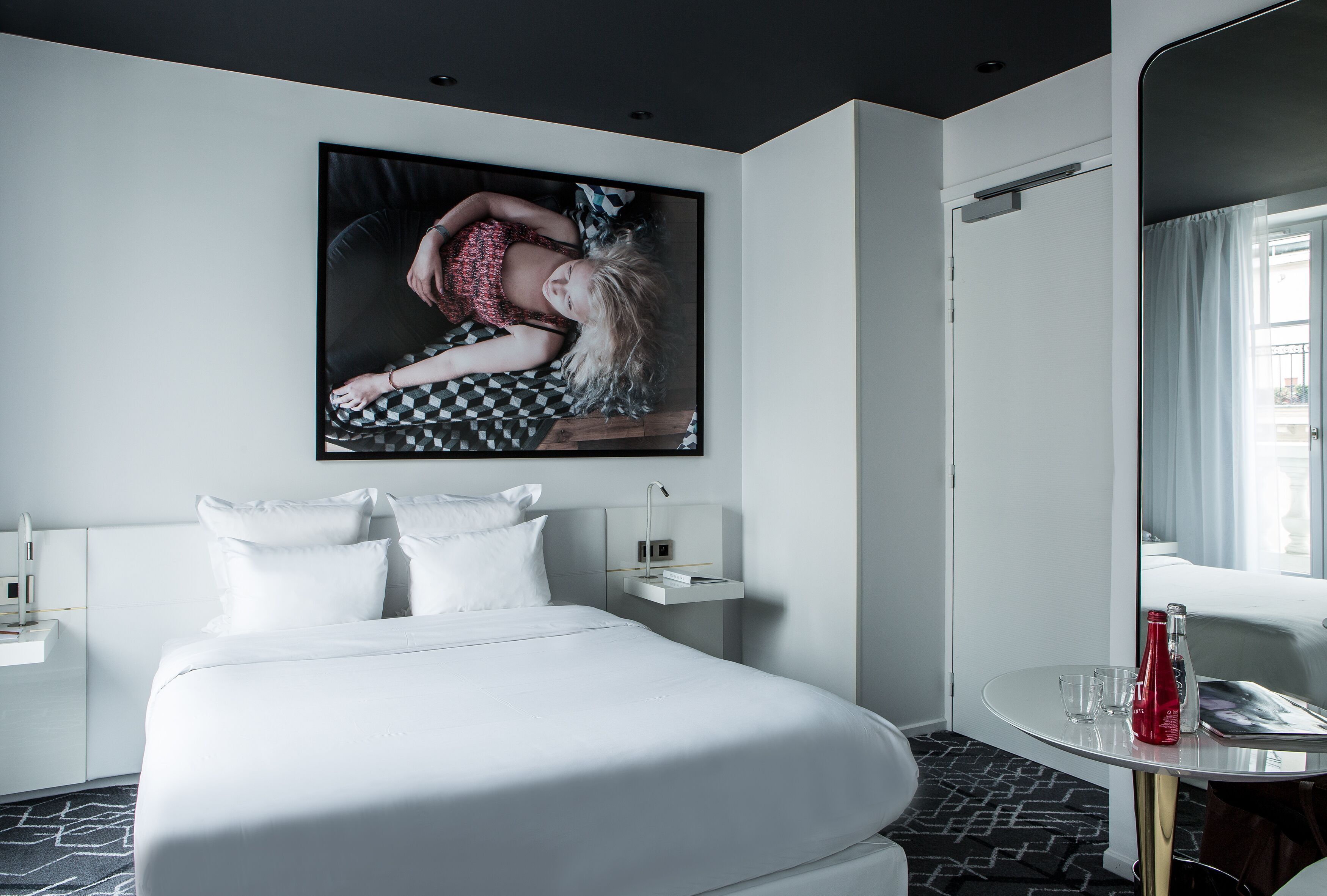 A modern hotel room with a large bed, a bedside table with a lamp, a small shelf, and a large mirror.