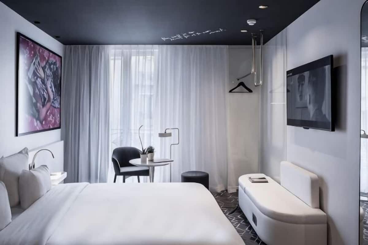A modern hotel room with a large bed, a small table with a chair, a sofa, a TV, and a painting on the wall.