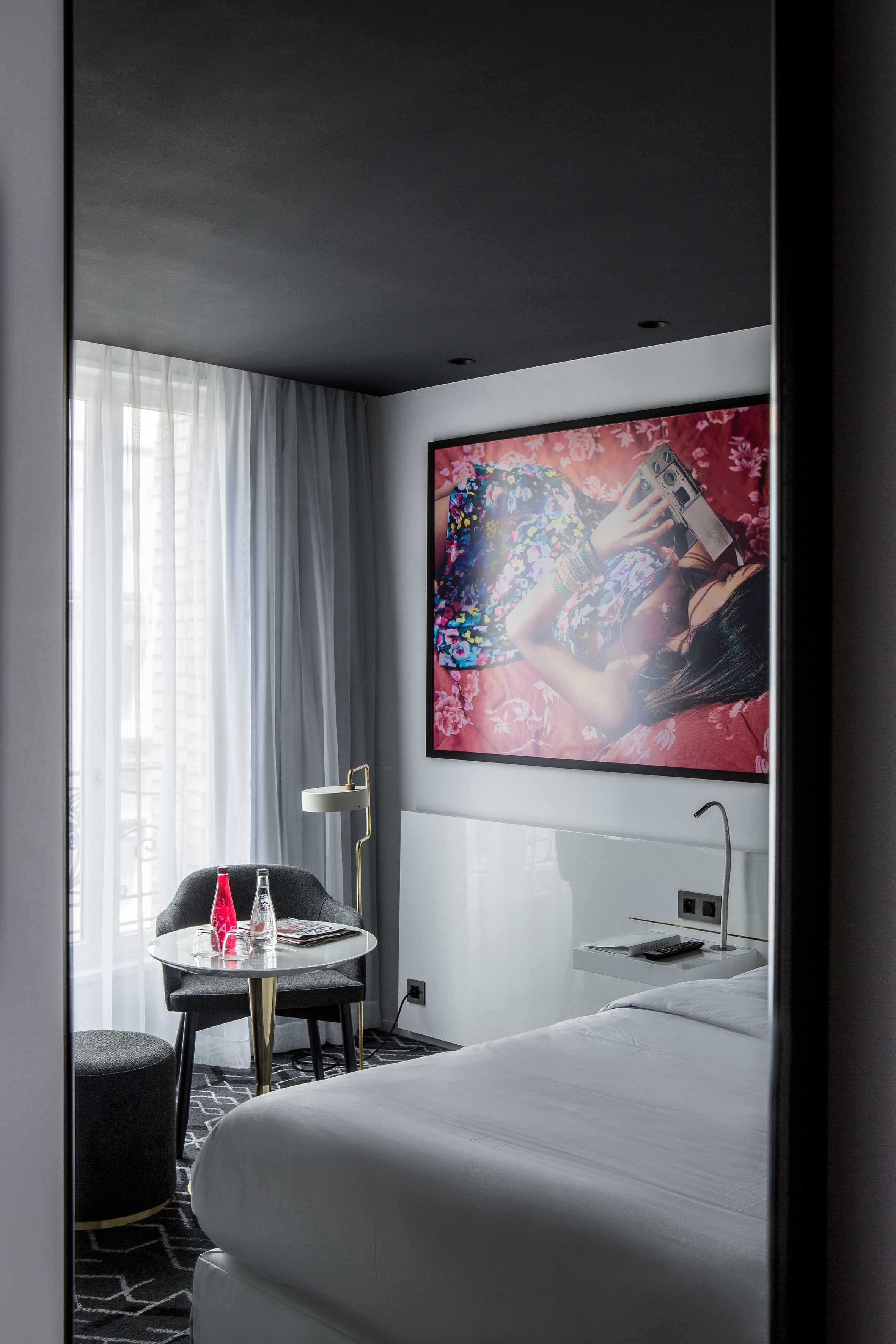 Una habitación de hotel moderna con una cama grande, una pequeña mesa redonda con un florero rojo, una silla, una mesita de noche con un control remoto y una obra de arte floral grande en la pared.