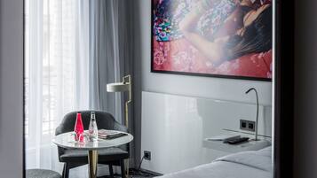 Una habitación de hotel moderna con una cama grande, una pequeña mesa redonda con un florero rojo, una silla, una mesita de noche con un control remoto y una obra de arte floral grande en la pared.