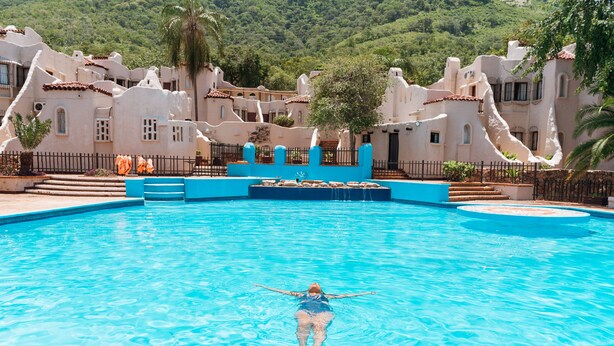 En swimmingpool med en person, der flyder i vandet, omgivet af en hvid bygning med terracotta-tag og et bjergrigt landskab i baggrunden.